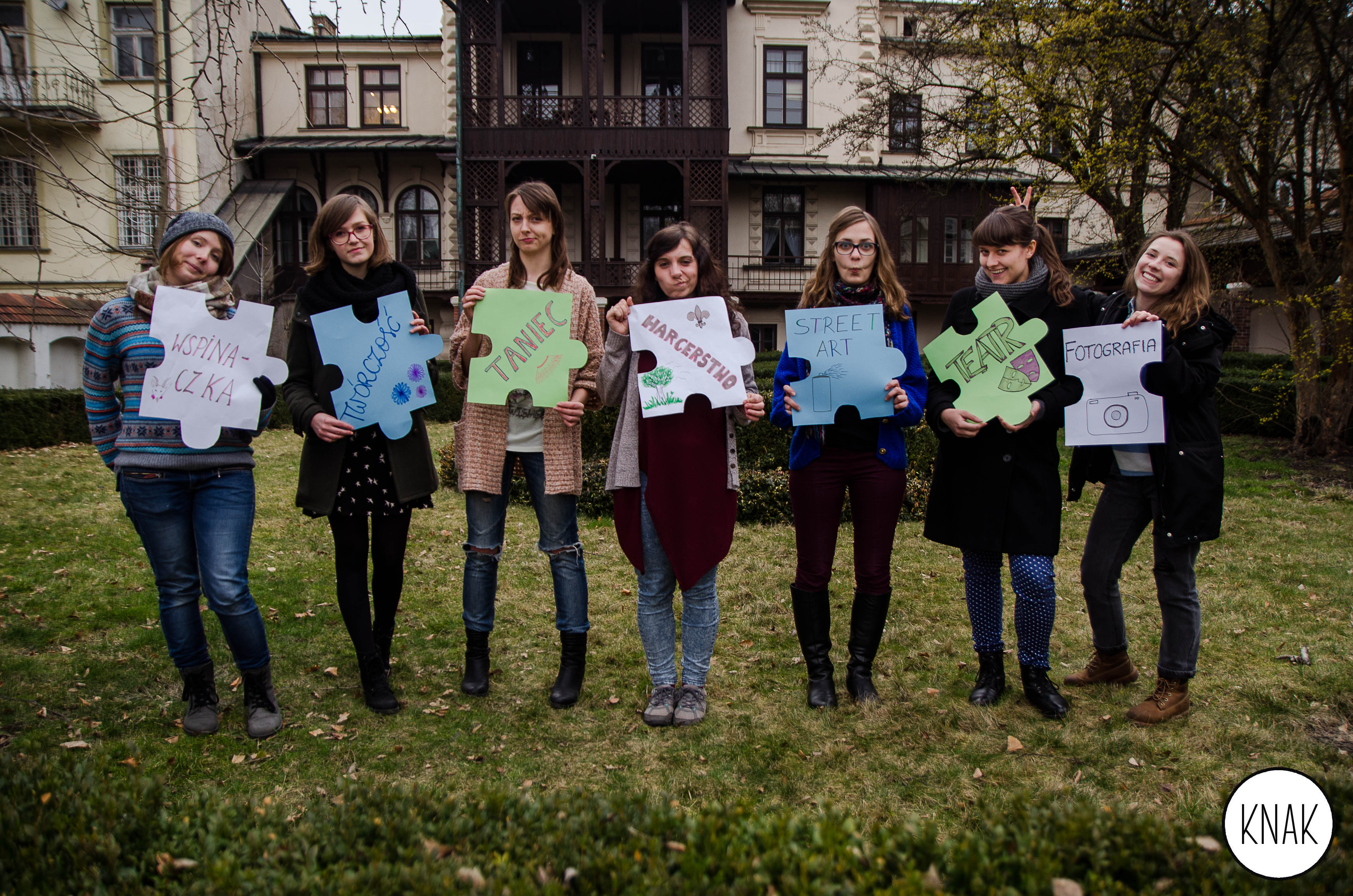 Zdjęcie członkow koła naukowego. Na zdjęciu siedem osób, trzymających transparenty z hasłami: "wspinaczka", "twórczość", "taniec", "harcerstwo", "streetart", "teatr", "fotografia".
